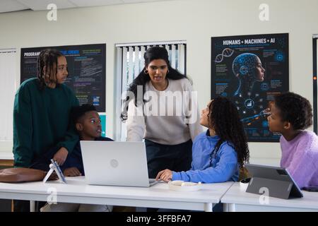 Diverse instructor with youth students leaning over silver laptop, teaching coding in classroom lab Stock Photo