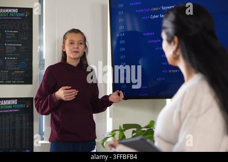 Female teenage student in hoodie pointing with marker at lab monitor showing code, teacher with pad Stock Photo