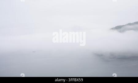 A minimalist landscape photograph featuring a dense, white fog heavily obscuring a body of water. On the right side, the dark silhouette of a forested Stock Photo