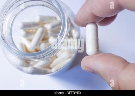 Hand Holding White Capsule Next to Open Glass Jar of Pills, Medication Concept Stock Photo