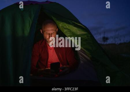 Focused male hiker sitting inside green camping tent using smartphone with serious expression during night bivouac in mountains staying connected in w Stock Photo