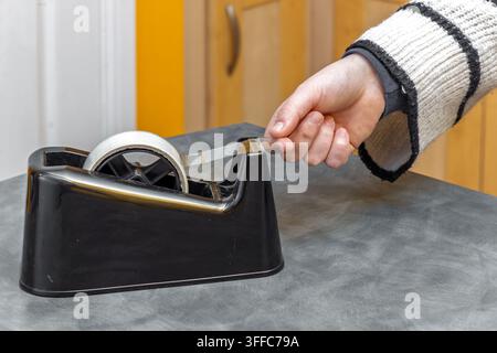 Close-up of hand and sleeve pulling clear adhesive tape from heavy black desktop dispenser sitting on grey surface, wooden cabinet visible in backgrou Stock Photo