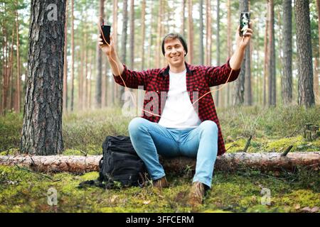 happy man in a forest holds his smartphone charging with a portable power bank. He stays connected while traveling outdoors, solving a low battery eme Stock Photo