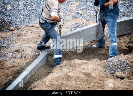 Two construction workers engaged in installation of concrete road curbing elements, carefully placing heavy blocks into prepared trench, ensuring prop Stock Photo