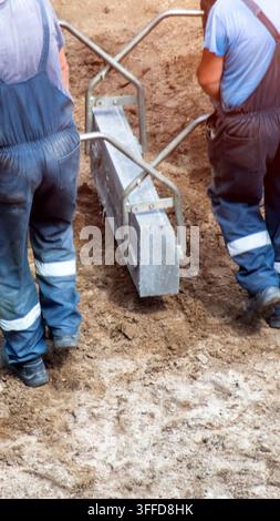 Construction workers busy with installation of road curb. Two people in work uniforms handling heavy concrete element with lifting device. Focus on te Stock Photo