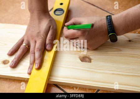 Man marks measurement line on pine board using pencil and level for precise woodworking cuts. Concept of precision planning and accurate measurement t Stock Photo