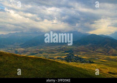 High-angle view captures a serene valley nestled between majestic mountains in Kyrgyzstan. Lush grassy slopes lead to a mosaic of agricultural fields and a small settlement. Stock Photo
