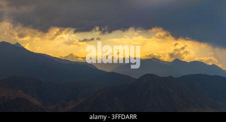 Panoramic view captures multiple mountain ranges silhouetted against a vibrant sky. Dark, textured slopes contrast with distant peaks illuminated by warm golden light breaking through heavy clouds. Stock Photo