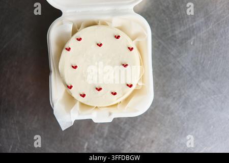Round white frosted cake with small red heart decorations in takeaway box on metal surface, with cake, frosting, white, heart detail, showing red Stock Photo