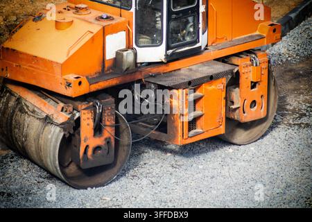 Close-up view of industrial asphalt paving roller working on road foundation. Large orange heavy equipment with steel drums compacting layer of gray c Stock Photo