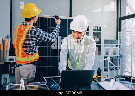 Two Asian engineers wearing hardhats and safety vests discussing solar panel installation and renewable energy project in the office. Stock Photo