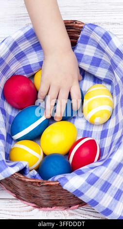 Child hand selecting blue Easter egg from collection of vibrant dyed eggs inside brown wicker basket. Eggs are red blue yellow with white stripes. Bas Stock Photo