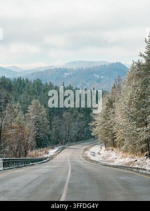 Winter road in snowy mountains. Autumn or wintertime in mountains. Asphalted road through fir tree forest and white snowy mountain peaks in the distance on background. Amazing landscape. Copy space Stock Photo