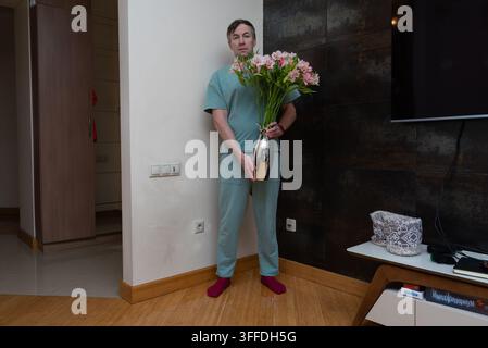 A 40-year-old man in green loungewear stands in a modern living room holding a silver vase filled with pink flowers. Stock Photo
