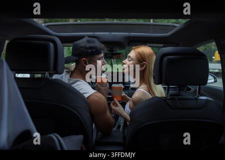 Back seat view of an intimate young couple holding takeaway coffee cups inside a car, leaning in close to each other. Driver and redhead passenger dur Stock Photo