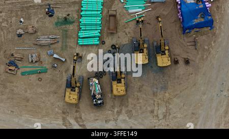 Aerial view of construction site featuring multiple yellow excavators, stacks of green pipes, and various building materials on dirt ground in Longmon Stock Photo