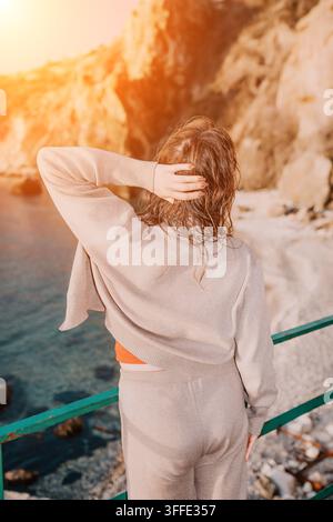 Woman beach sunset view from behind of a young lady in beige clothing looking at the ocean and cliffs during golden hour Stock Photo