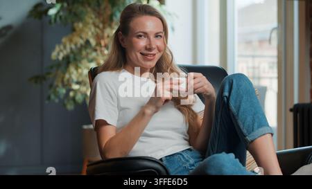 Happy Caucasian middle-aged woman relaxing looking at camera smiling enjoying tasty coffee at morning at home adult female relax drink tea beverage Stock Photo