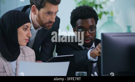 Business team concentrated three multiethnic employers comparing documents stats and computer data talking in office. Caucasian leader with African Stock Photo