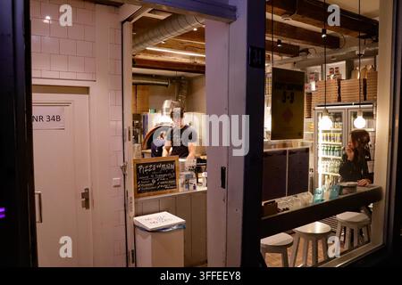 A kitchen worker is preparing a pizza in a food outlet while a customer sat on a stool looks on. A drinks refrigerator is visible in the background. Stock Photo