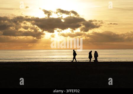 A dramatic sunset with the sun's rays breaking through the clouds over a calm sea. Three figures are walking along the beach. Stock Photo