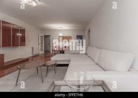 Living room with white sectional sofa, oval coffee tables, glass dining set, and view into hallway. Stock Photo