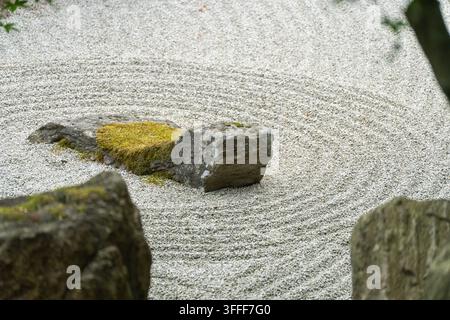 A moss-topped stone rests amid concentric raked gravel circles in a zen garden, flanked by two other large rocks in close-up view. Stock Photo