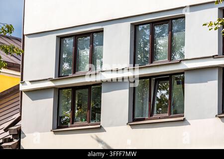Low-angle exterior shot of a modern light-grey building facade featuring neat rows of dark-framed windows reflecting autumn green leaves. Clean archit Stock Photo