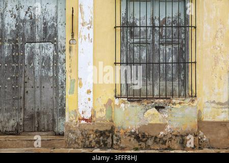 View of a weathered yellow colonial wall with a rustic wooden door and a window protected by iron bars in Remedios, Villa Clara, Cuba. Stock Photo