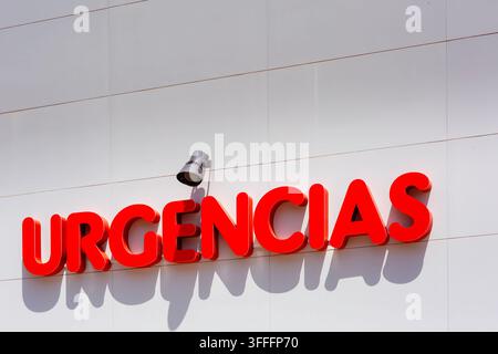 Red three dimensional letters spelling urgencias are mounted on a white tiled hospital exterior wall. A grey spotlight fixture sits above the sign. Br Stock Photo