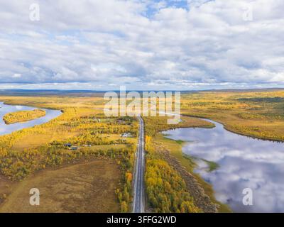 Aerial shot of an arctic road leading through golden autumn tundra near traditional cabins and a mirrored river in northern Sweden Stock Photo