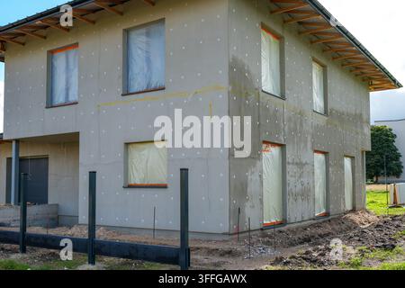 Two-story residential house under construction with insulated exterior walls, protected windows, exposed wiring and unfinished roof eaves and grounds Stock Photo