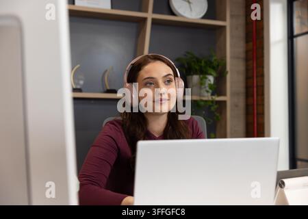 Adult female office worker sitting at desk in office, using silver laptop with pink headphones Stock Photo