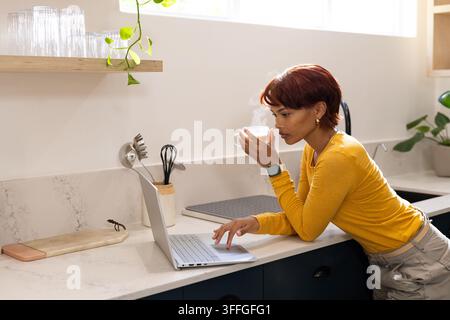 African American woman leaning on counter in kitchen, holding steaming cup, using laptop Stock Photo