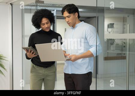 Asian, African-American male team reviewing silver laptop, dark tablet near glass wall in corridor Stock Photo
