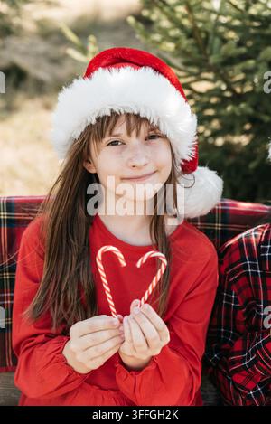 Christmas in July with smiling girl wearing Santa hat and holding heart made from candy canes in sunny summer garden beside evergreen Christmas tree. Stock Photo