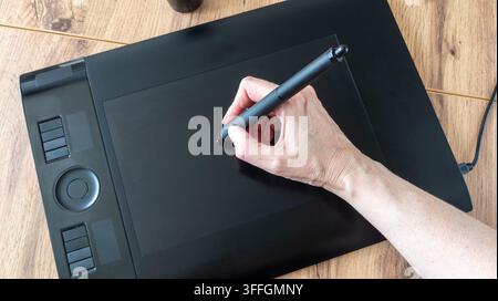 Hand holds a digital stylus and draws on a black graphics tablet. Creative process of creating a digital design on a wooden table.  Flat lay. Stock Photo