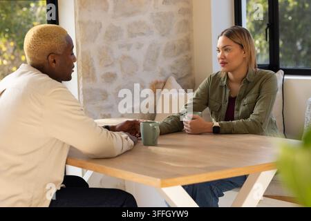 Diverse couple sitting in home dining nook at light wood table holding ceramic mugs Stock Photo