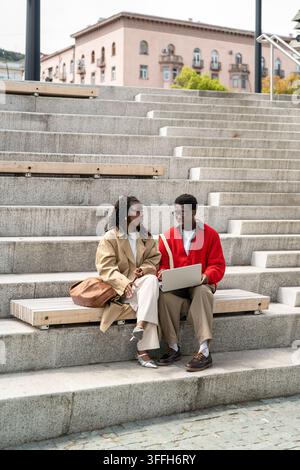 Stylish Black student friends discussing university work with laptop on outdoor steps together Stock Photo