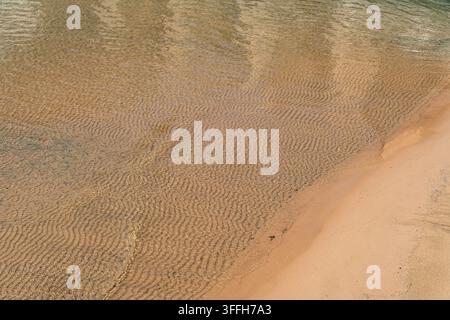 Close-up texture of golden sandy beach and crystal-clear water, creating a natural summer background with warm colors and serene coastal atmosphere Stock Photo