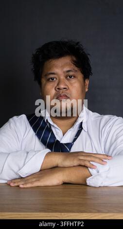 A serious young Asian male professional sits directly facing the camera with folded arms resting on a wooden desk against a pitch black backdrop. The Stock Photo