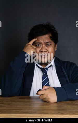A thoughtful young Asian male executive sits at a wooden desk, pressing one index finger against the temple in a classic gesture of deep concentration Stock Photo