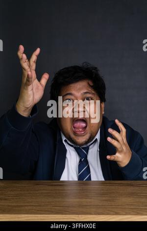 A terrified young Asian male employee screams in extreme panic with both hands raised dramatically in the air above a wooden desk. Set against a stark Stock Photo