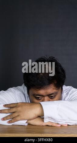 A young Asian male office employee rests the head completely face down on crossed arms over a smooth wooden desk against a solid dark gray wall. The e Stock Photo