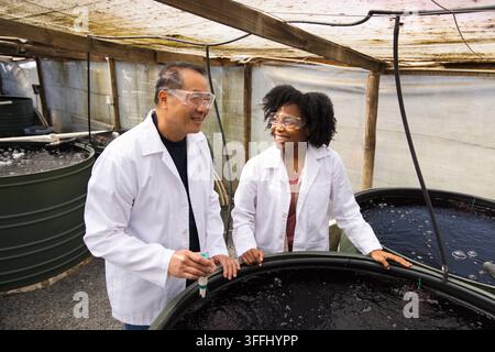 Diverse researchers examining round hatchery tank in greenhouse holding probe and wearing lab coats Stock Photo