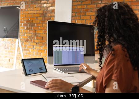 African American woman wearing top at white desk using laptop spreadsheet, tablet charts Stock Photo