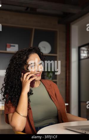 African American woman sitting at round white table in office, holding smartphone, typing on laptop Stock Photo