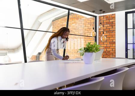 African American woman leaning over conference table, holding phone and writing notes in turtleneck Stock Photo
