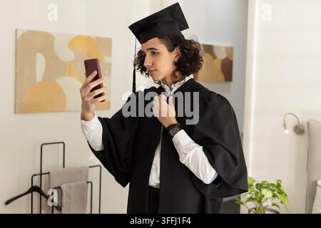 Man holding red smartphone and adjusting black graduation gown in bedroom with plant Stock Photo