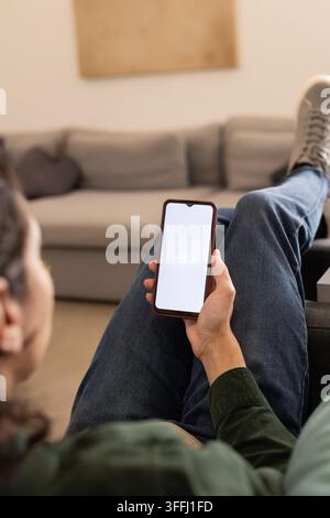 African American man reclining on sofa holding phone with blank screen at home wearing denim jeans Stock Photo